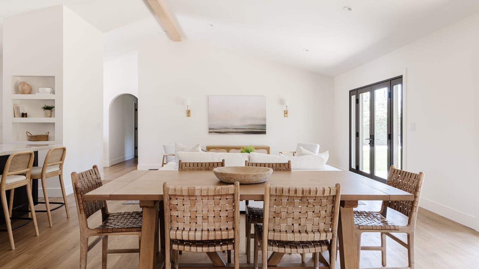 Open dining room with a long oak table, woven leather chairs, a vaulted beamed ceiling, and black steel doors opening to the garden