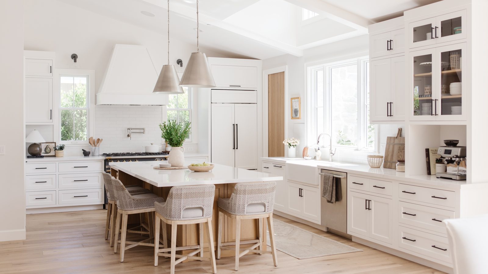 Bright white kitchen with a skylit island, custom plaster hood, woven counter stools, and warm oak flooring