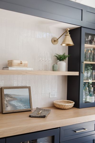 Butler's pantry bar with navy cabinetry, a floating oak shelf, brass sconce, glass-front uppers and framed art
