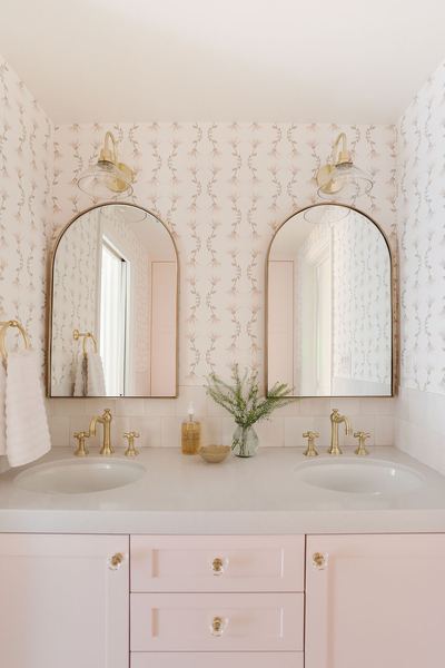 Bathroom with blush pink cabinetry, two arched brass mirrors, brass faucets and delicate patterned wallpaper