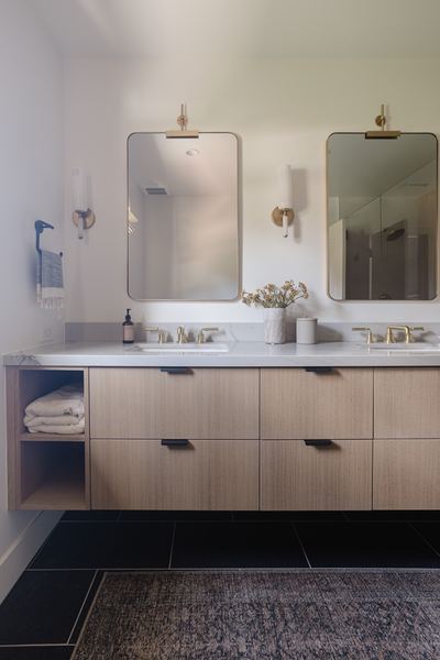 Double vanity bathroom with white oak cabinetry, twin arched mirrors, brass sconces and dark stone tile flooring