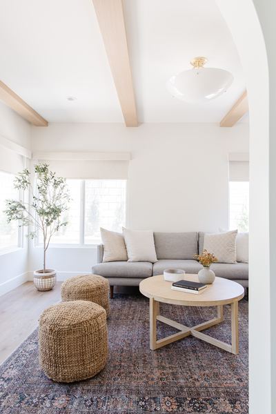 Living room with a light grey sectional, woven poufs, round oak coffee table and an olive tree beneath white-painted ceiling beams