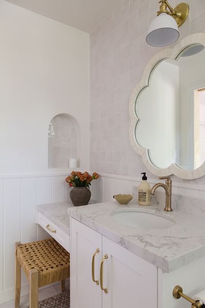 Powder room with a scalloped mirror, marble counter, brass faucet, arched wall niche and a woven bench