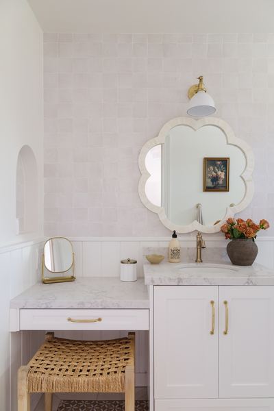 Powder room with a scalloped mirror, marble countertop, woven stool, brass faucet and a vase of orange ranunculus
