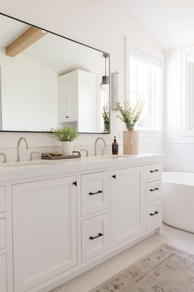 Primary bathroom with white cabinetry, black hardware, a freestanding tub beneath a window and an exposed ceiling beam