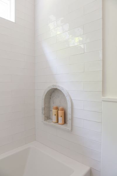 Shower surround in stacked white gloss tile with an arched recessed niche framed in veined marble, lit by daylight from a high window above a white tub