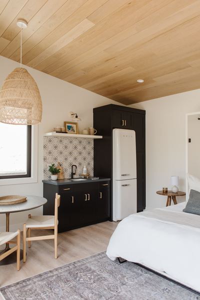 Guest suite with a warm wood plank ceiling, woven pendant light, black kitchenette cabinetry and a pale upholstered bed