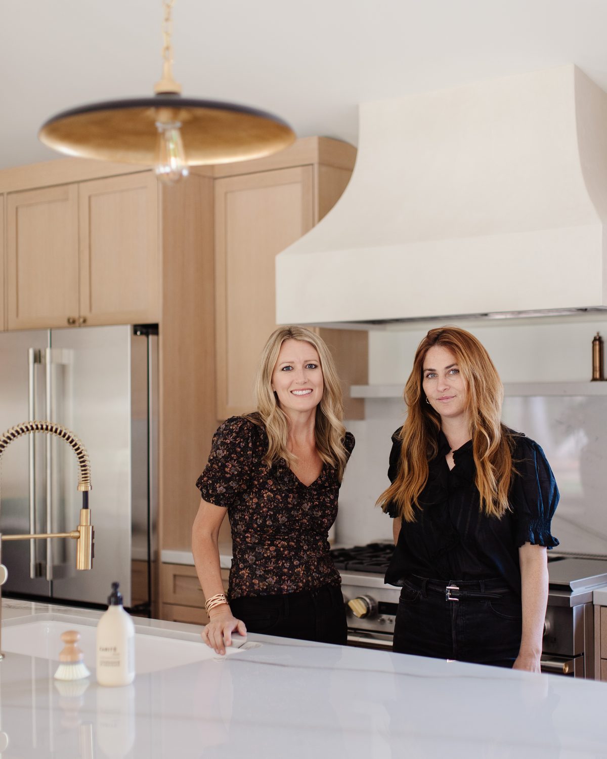 The Flourish Home Studio design team in a completed kitchen with white oak cabinetry, a plaster range hood, and a brass pendant light